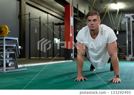 Man doing push-up on padded gym floor wearing sportswear and smartwatch with barbell rack behind Man doing push-up on padded gym floor wearing sportswear and smartwatch with barbell rack behind 133292493