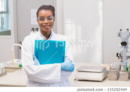 African American woman wearing lab coat and gloves holding blue binder in lab with microscopes African American woman wearing lab coat and gloves holding blue binder in lab with microscopes 133292494