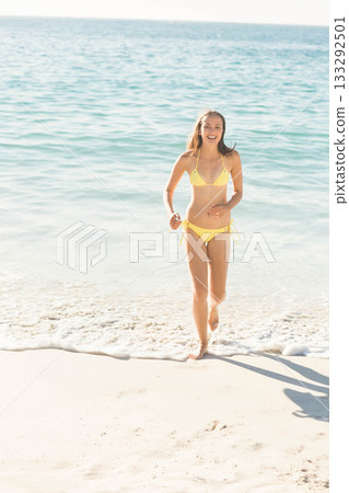 Woman wearing bright yellow bikini running from ocean waves onto white sandy beach under clear sky Woman wearing bright yellow bikini running from ocean waves onto white sandy beach under clear sky 133292501