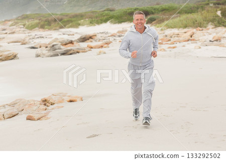Adult man running along sandy shoreline wearing light grey hoodie grey sweatpants and running shoes Adult man running along sandy shoreline wearing light grey hoodie grey sweatpants and running shoes 133292502