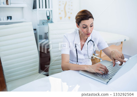 Mid adult medical professional wearing stethoscope and using laptop in exam room, copy space 133292504
