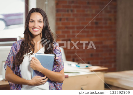Woman standing in office holding tablet next to wooden desk cluttered with stacks papers notebooks Woman standing in office holding tablet next to wooden desk cluttered with stacks papers notebooks 133292509
