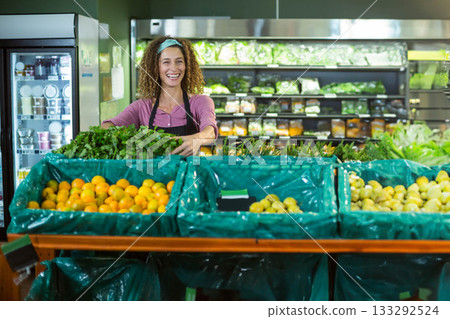 Female grocery clerk wearing apron and headband arranging leafy greens in bins at produce section Female grocery clerk wearing apron and headband arranging leafy greens in bins at produce section 133292524