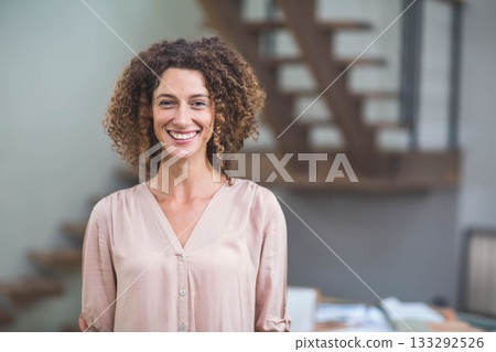 Woman standing in office wearing pink blouse by wooden staircase and desk with books and papers Woman standing in office wearing pink blouse by wooden staircase and desk with books and papers 133292526