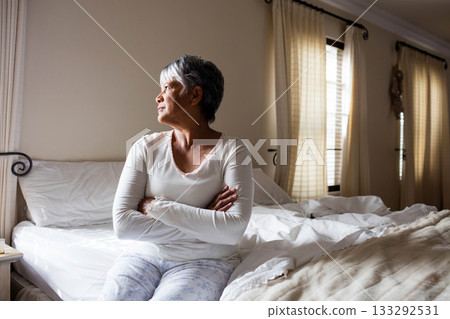 Senior woman sitting on unmade bed in bedroom crossing arms and gazing through curtains, copy space 133292531