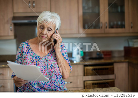 Senior woman talking on smartphone while reading paper in home kitchen near coffee maker 133292539