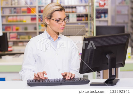 Female pharmacist in lab coat typing on keyboard at pharmacy counter with medication boxes, bottles Female pharmacist in lab coat typing on keyboard at pharmacy counter with medication boxes, bottles 133292541