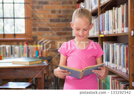 Female child holding open book and reading pages in library aisle beside wooden table with books 133292542