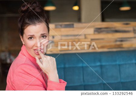 Teenage female wearing blazer biting finger on bench by slat wall in cafe with pendant lights Teenage female wearing blazer biting finger on bench by slat wall in cafe with pendant lights 133292546