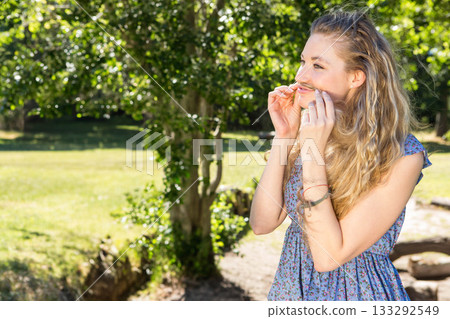 Woman standing in park holding grass blade under nose wearing bracelets, looking right, copy space 133292549