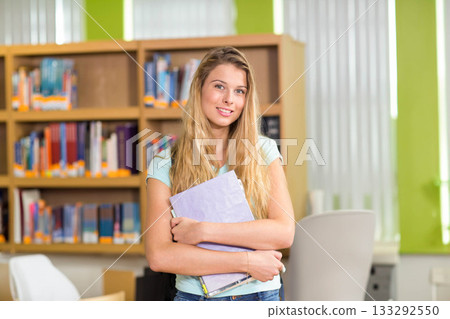 Teenage girl standing inside library holding light purple binder and notebooks near bookshelves Teenage girl standing inside library holding light purple binder and notebooks near bookshelves 133292550