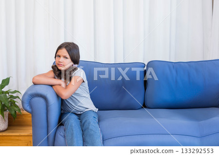 Female child sitting on blue couch in living room with potted plant, white curtains, copy space 133292553