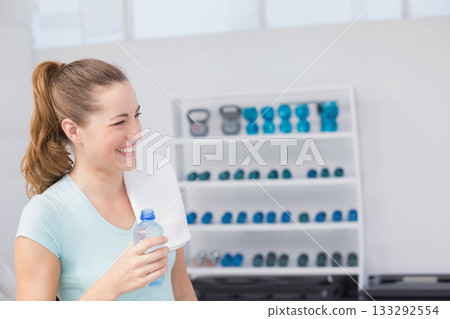 Woman holding towel and water bottle while standing by weight rack in fitness studio, copy space 133292554