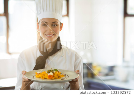 Female chef wearing pleated hat holding plate of pasta with herbs and cheese in bright kitchen 133292572