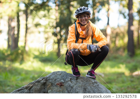 Woman crouching on rock in forest wearing orange jacket bicycle helmet and backpack 133292578