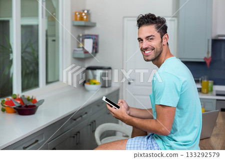 Man sitting on stool at kitchen counter holding smartphone and laptop with fruit bowl, copy space 133292579
