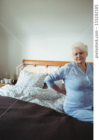 Senior woman sitting on bed in bedroom wearing pajamas supporting back by nightstand, copy space Senior woman sitting on bed in bedroom wearing pajamas supporting back by nightstand, copy space 133292585