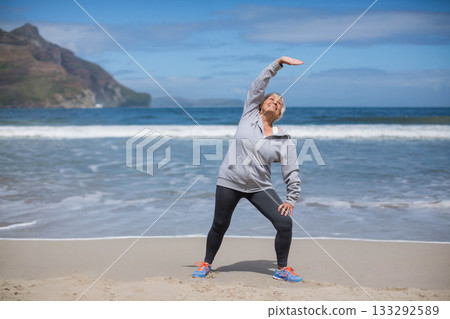 Senior woman stretching torso toward left on sandy beach wearing sportswear and athletic shoes Senior woman stretching torso toward left on sandy beach wearing sportswear and athletic shoes 133292589