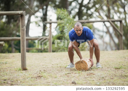 African American man in workout gear lifting wooden log on grass field near rail fence 133292595