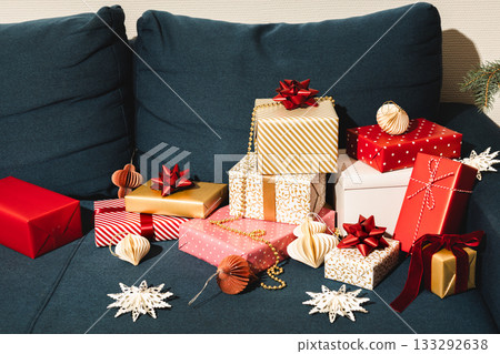 Holiday scene with wrapped Christmas presents and festive decorations on dark blue sofa, red and gold gift boxes with bows, cozy winter celebration atmosphere, modern home interior style Holiday scene with wrapped Christmas presents and festive decorations on dark blue sofa, red and gold gift boxes with bows, cozy winter celebration atmosphere, modern home interior style 133292638