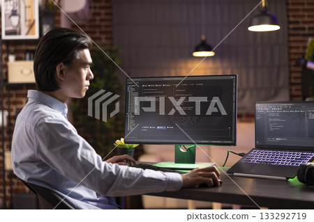 Self employed man sitting indoors during evening hours, concentrating on multiple screens, working on html codes. Male asian programmer reviewing online applications to develop new website ideas. Self employed man sitting indoors during evening hours, concentrating on multiple screens, working on html codes. Male asian programmer reviewing online applications to develop new website ideas. 133292719