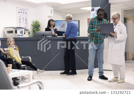Elderly female physician in lab coat consults with male patient in hospital waiting room. Senior doctor with laptop discusses recovery plan with black man as receptionist assists clinic visitors. 133292721