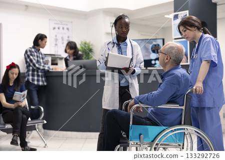 Senior man with disability attends medical consultation at hospital, speaking with doctor in waiting room. Female physician listens to wheelchair patient, taking notes on clipboard in clinic lobby. 133292726