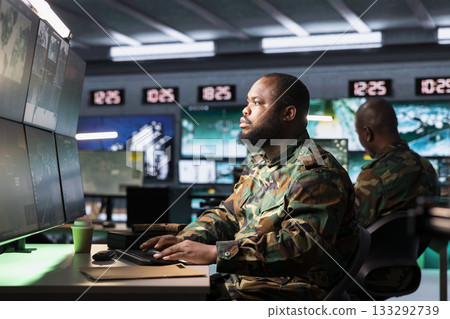 Soldier in war room analyzing aerial reconnaissance data to come up with warfare winning tactics. African american intelligence agency agent controlling drones attacking strategic targets Soldier in war room analyzing aerial reconnaissance data to come up with warfare winning tactics. African american intelligence agency agent controlling drones attacking strategic targets 133292739