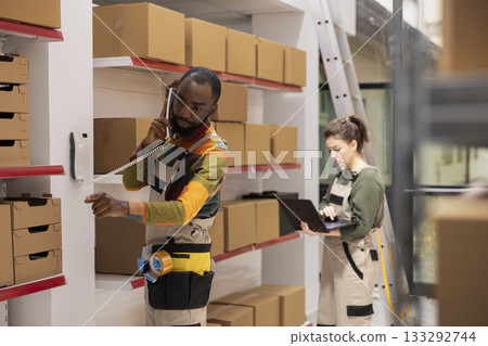 Black male staff member in coveralls manages phone calls about custom orders, ensuring dispatch accuracy, shipment processing and logistics for e-business small delivery exports. Black male staff member in coveralls manages phone calls about custom orders, ensuring dispatch accuracy, shipment processing and logistics for e-business small delivery exports. 133292744