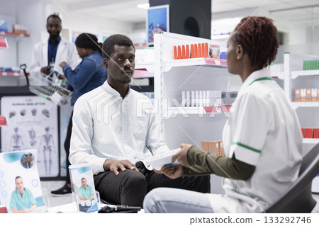 Black healthcare assistant offers personalized medication advice to a young customer in a modern stacked pharmacy shop, recommending vitamins and drugs for effective self care. Black healthcare assistant offers personalized medication advice to a young customer in a modern stacked pharmacy shop, recommending vitamins and drugs for effective self care. 133292746