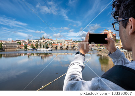 Prague, Czech Republic, August 9, 2023. A young Caucasian man takes photos with his cell phone of the enchanting view of the castle and cathedral dominating the hill. A lifestyle using technology. Prague, Czech Republic, August 9, 2023. A young Caucasian man takes photos with his cell phone of the enchanting view of the castle and cathedral dominating the hill. A lifestyle using technology. 133293007