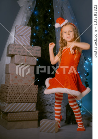 Young girl in a red Santa dress dancing joyfully near a mountain of Christmas presents by a beautifully decorated tree. Young girl in a red Santa dress dancing joyfully near a mountain of Christmas presents by a beautifully decorated tree. 133293822
