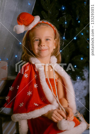Girl dressed as a red elf with a soft cape sitting on a sled near a Christmas tree decorated with blue lights. 133293851