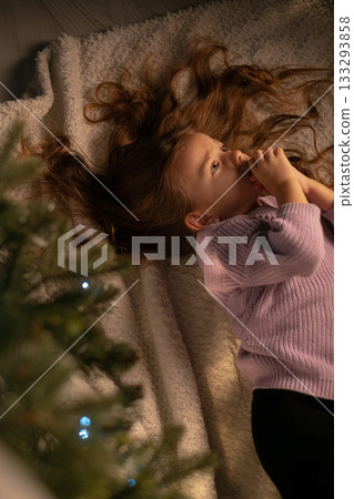 Girl in festive sweater lying under Christmas tree enjoying cozy holiday atmosphere and magic of celebration 133293858