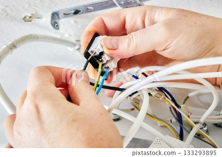 Mounting process involves technician joining electrical wires to terminal block of LED light fitting on ceiling surface in countryside home, device partially open. Mounting process involves technician joining electrical wires to terminal block of LED light fitting on ceiling surface in countryside home, device partially open. 133293895
