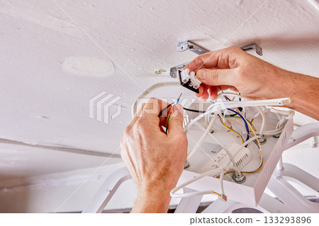 Wiring is being connected by electrician during installation of new LED chandelier on white ceiling in rural house, wires and fixture visible in frame. Wiring is being connected by electrician during installation of new LED chandelier on white ceiling in rural house, wires and fixture visible in frame. 133293896