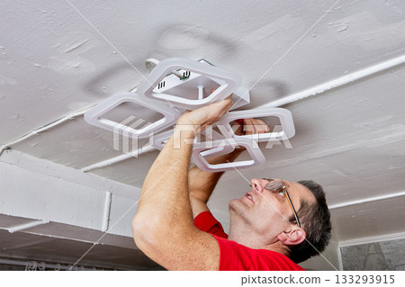 Lighting installation work includes electrician fitting LED chandelier to ceiling in rural house during setup of new lighting fixture. 133293915