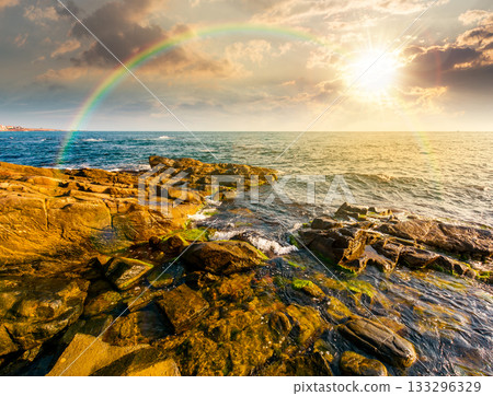 sunset at the rocky sea beach. beautiful view of coast landscape in summer. sun over horizon and cloudy sky. dramatic scenery with rock formations under rainbow. great seascape for weekend background 133296329