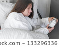 A woman, wrapped in a soft robe, with a wary expression on her face, looking at the alarm clock, among light pillows and a calm interior, anticipating the silence of the early morning. Copy space 133296336