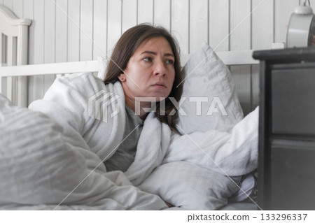 A woman, wrapped in a soft robe, with a wary expression on her face, looking at the alarm clock, among light pillows and a calm interior, anticipating the silence of the early morning. Copy space 133296337