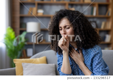 Woman with curly hair suffering from a painful cough, feeling unwell due to a cold, flu, allergy, or other respiratory illness while resting on a sofa in her home 133296517