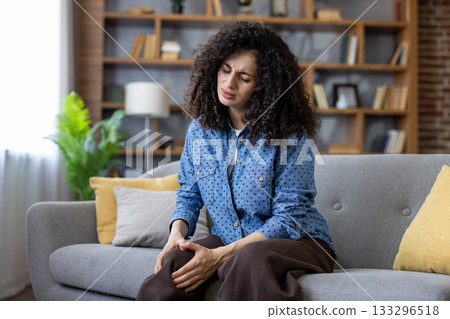 Woman sitting on sofa at home clutching her knee in acute pain, showing joint discomfort and possible arthritis or injury, appearing unhappy and strained in a living room setting Woman sitting on sofa at home clutching her knee in acute pain, showing joint discomfort and possible arthritis or injury, appearing unhappy and strained in a living room setting 133296518