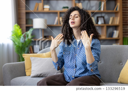 Woman feeling overheated and uncomfortable during a heatwave, fanning herself with hands while sitting on a sofa in a modern apartment, showing distress from high summer temperatures Woman feeling overheated and uncomfortable during a heatwave, fanning herself with hands while sitting on a sofa in a modern apartment, showing distress from high summer temperatures 133296519