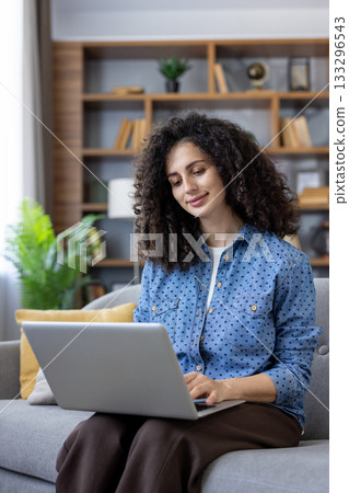 Woman with curly hair sitting on a cozy sofa, smiling and focused while typing on a laptop, engaging in remote work or online communication from her modern living room 133296543