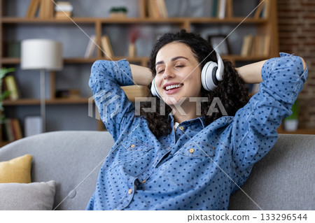 Young woman with closed eyes and a happy expression enjoying music through white headphones while sitting comfortably on a gray sofa in her home, embracing leisure and calm 133296544