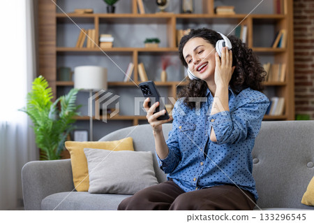 Woman with curly hair listening to music through headphones, holding smartphone, smiling and relaxing on a comfortable sofa in her living room at home 133296545