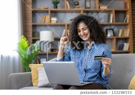 Young woman triumphantly purchasing goods online with a credit card, using a laptop, happily sitting on a couch in her living room, celebrating a successful transaction Young woman triumphantly purchasing goods online with a credit card, using a laptop, happily sitting on a couch in her living room, celebrating a successful transaction 133296553