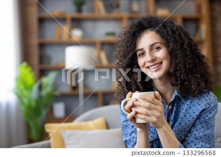 Smiling woman with curly brown hair relaxing on a cozy sofa in her modern living room, holding a warm mug and enjoying a peaceful morning break, content and comfortable Smiling woman with curly brown hair relaxing on a cozy sofa in her modern living room, holding a warm mug and enjoying a peaceful morning break, content and comfortable 133296568