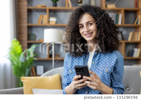 Young woman with curly hair sitting on a sofa, happily browsing her smartphone and looking at the camera, enjoying digital communication and connectivity in a cozy home environment Young woman with curly hair sitting on a sofa, happily browsing her smartphone and looking at the camera, enjoying digital communication and connectivity in a cozy home environment 133296598