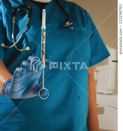 The dentist, in a blue lab coat and gloves, holds a carpal syringe in front of him, 133296763